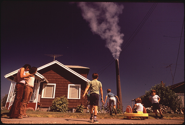 Children playing in their front yard as a smelter stack spews pollution over them.